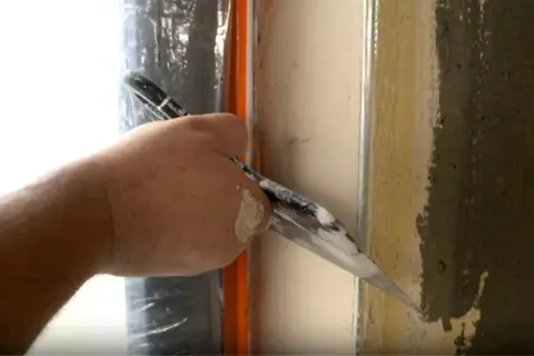 Ręka z kielnią wygładza tynk przy oknie, by ukryć krzywe glify po wstawieniu nowego okna.