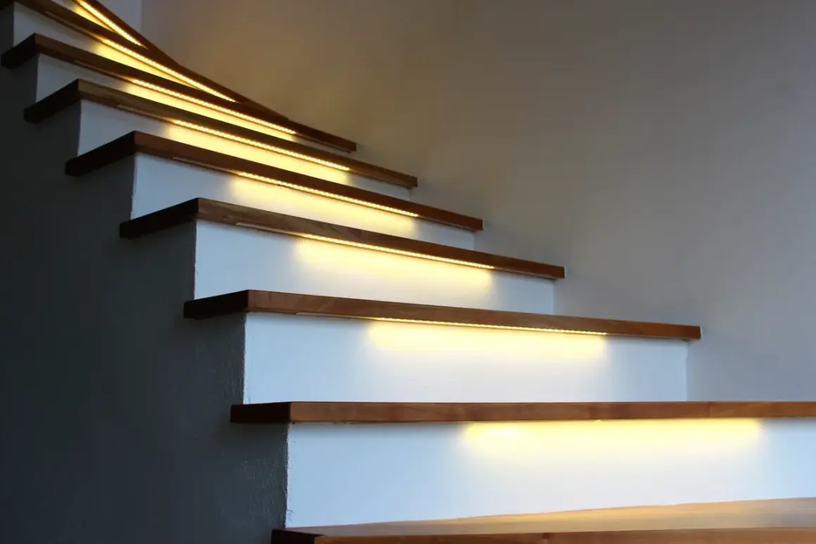 Oświetlenie LED nad klatką schodową z wysokim sufitem: taśmy LED zamontowane pod stopniami tworzą ciepłą, nowoczesną atmosferę.