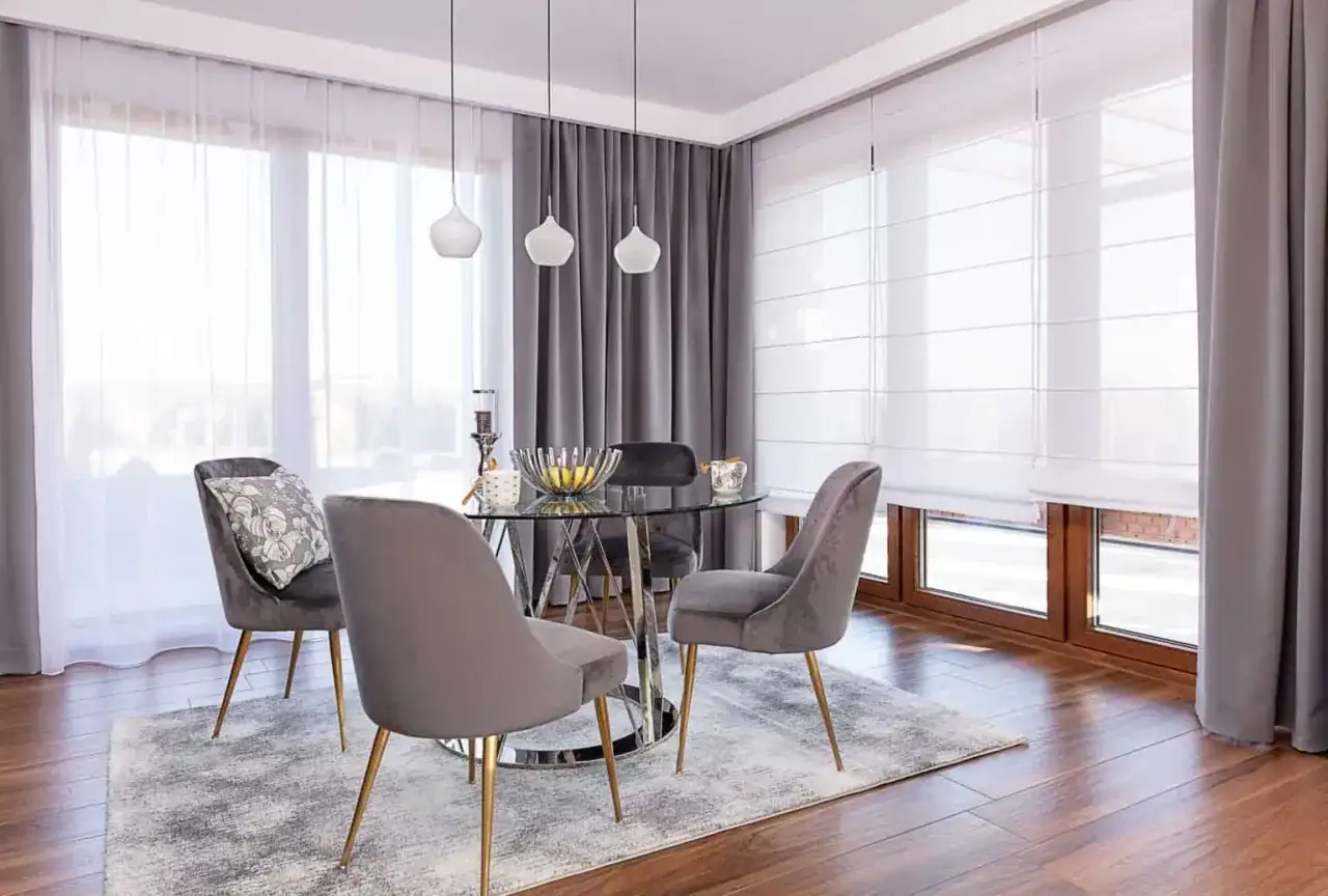 Elegancki salon z okrągłym stołem i welurowymi krzesłami. Zastanawiasz się, czy rolety rzymskie czy zasłony lepiej sprawdzą się w salonie? Tutaj widzisz połączenie obu rozwiązań.