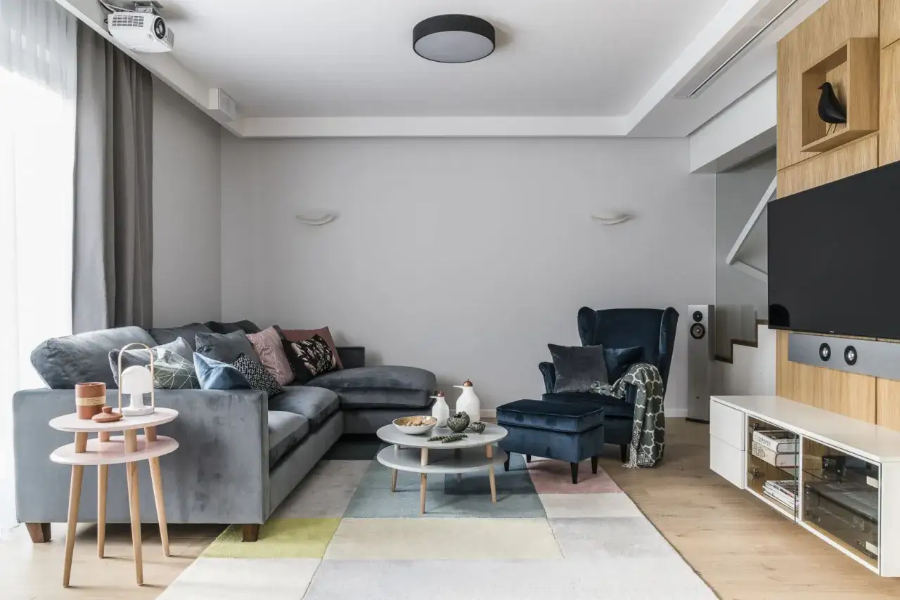 Jak ocieplić szary salon za pomocą dodatków i tekstyliów: miękki welurowy narożnik, kolorowy dywan i stylowe poduszki tworzą przytulną atmosferę.