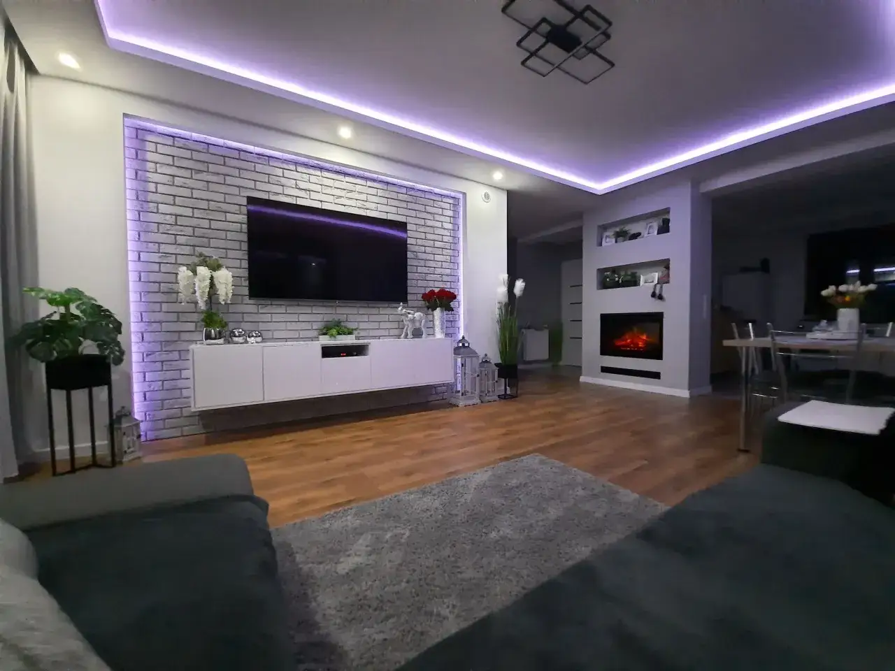 Nowoczesny salon z telewizorem na tle ceglanej ściany. Fioletowe oświetlenie LED za telewizorem i na suficie tworzy nastrojową atmosferę. Czy to oświetlenie LED za telewizorem nie męczy wzroku?