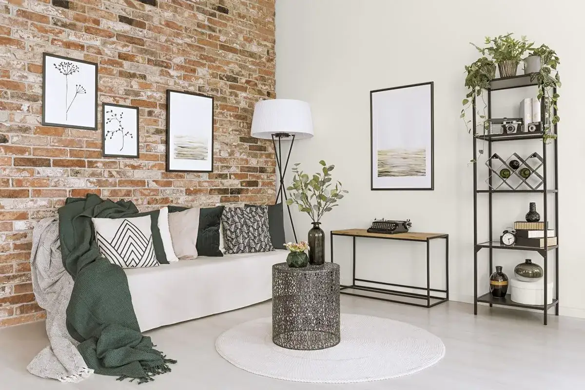 Nowoczesny salon z ceglaną ścianą. Jasna kanapa, zielone i wzorzyste poduszki, pledy. Stolik kawowy, lampa, regał z dekoracjami. Jak uniknąć efektu pomniejszenia pokoju?